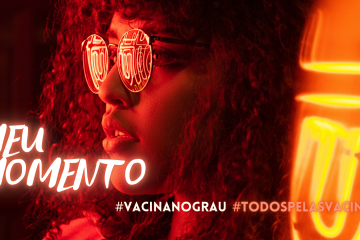 Uma jovens de perfil, com cabelos cacheados e óculos escuros, iluminadas por luzes neon laranjas com os escritos "meu momento" #vacinanograu #todospelasvacinas