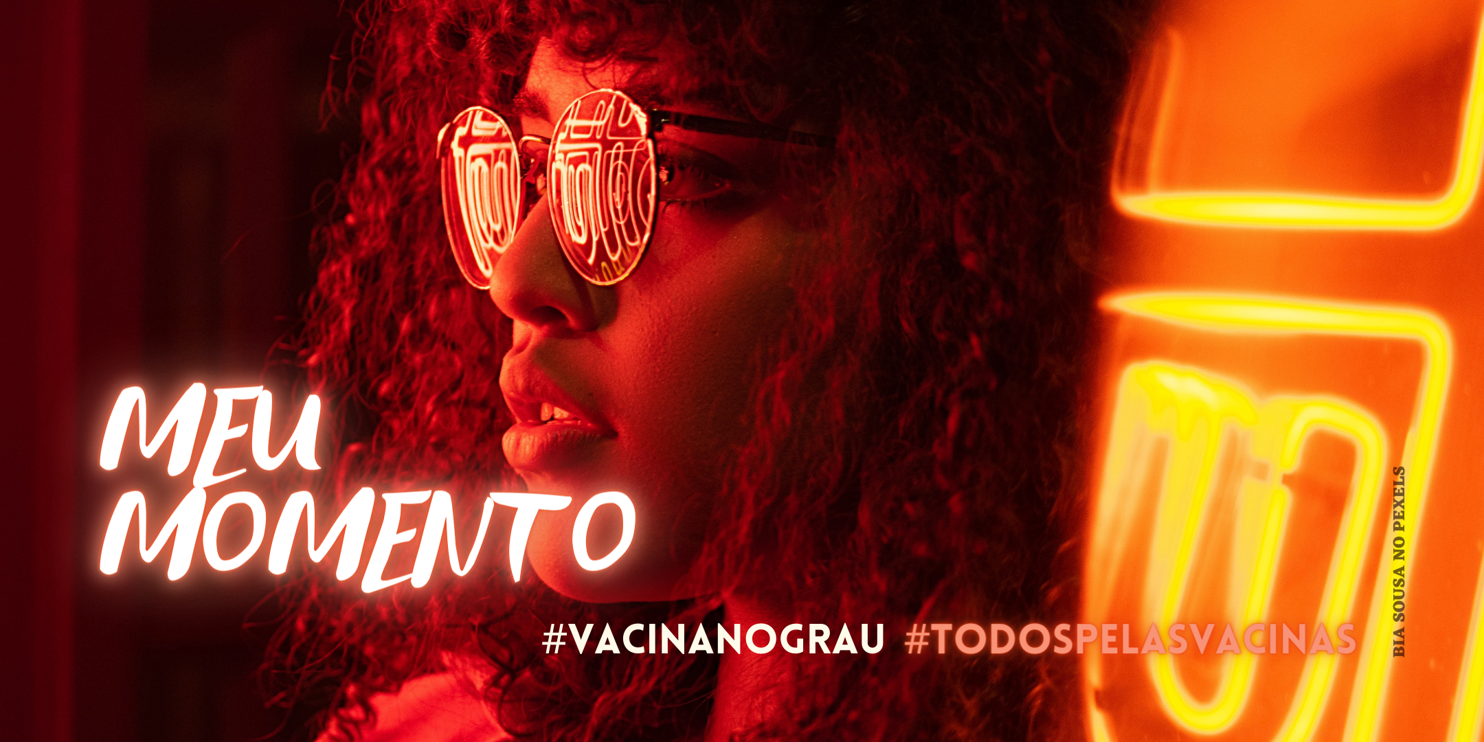 Uma jovens de perfil, com cabelos cacheados e óculos escuros, iluminadas por luzes neon laranjas com os escritos "meu momento" #vacinanograu #todospelasvacinas