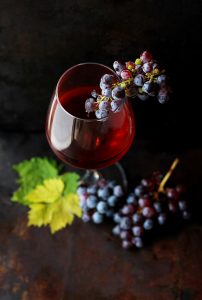 Taça com vinho dentro e um cacho de uva pendurado nela. Dois cachos de uva no pé da taça.