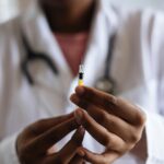 A imagem mosta um enfermeiro negro segurando com duas mãos uma seringa com líquido que se assemelha a vacina da tuberculose
