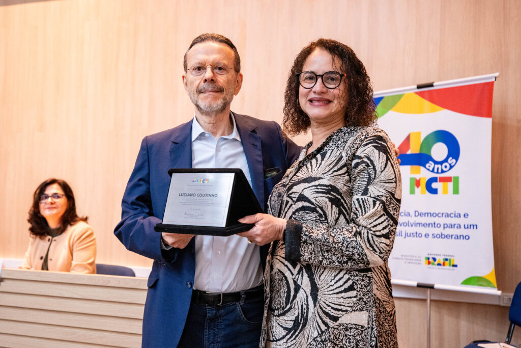 Foto do prof. dr. Luciano Coutinho e da ministra Luciana Santos. Ambos se abraçam de lado enquanto Coutinho e Luciana seguram a placa em homenagem ao professor entregue pela ministra. Os dois estão de pé e olham diretamente para a foto. Ao fundo, a professora Janaina observa os dois enquanto sorri.