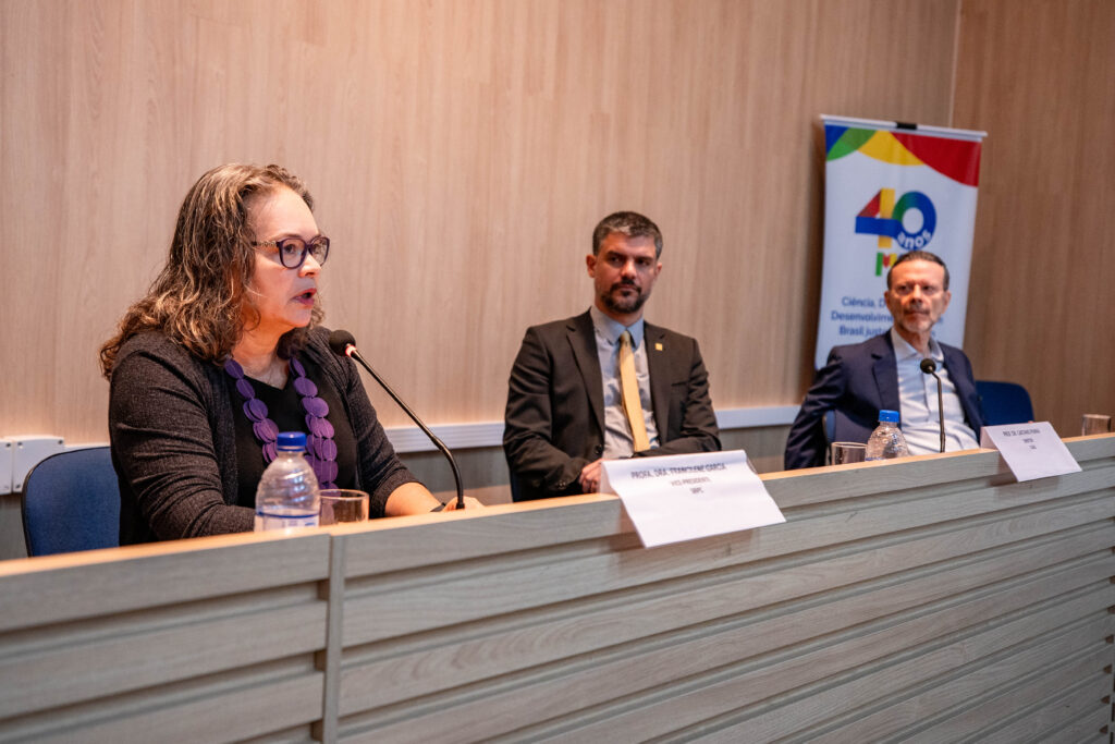 Foto de três pessoas sentadas à mesa. No primeiro plano, a prof. dra. Francilene Garcia, seguida de Caetano Penna e do prof. dr. Luciano Coutinho. Francilene olha para sua frente, enquanto os outros dois homens a observam.