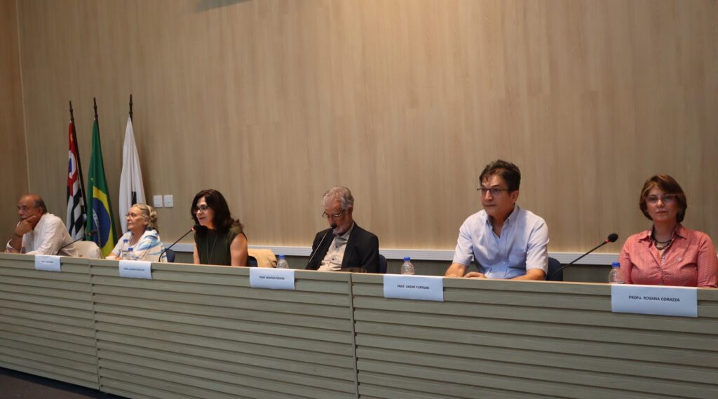 Foto de seis pessoas sentadas à mesa. Da esquerda para a direita da imagem: prof. dr. Roberto Dagnino, Profa. Dra. Leda Gitahy, Prof. Dr. Newton Pereira, Prof. Dr. André Furtado e prof. dra. Rosana Corazza. Todos olham para a frente enquanto Janaina fala.