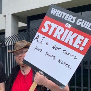 Trabalhador da indústria cinematográfica carrega um cartaz com os dizeres "Associação dos Escritores em Greve! A IA não vai levar em conta suas anotações bestas", ridicularizando as propostas dos estúdios de cinema para substituir escritores por inteligência artificial. Fonte: Wikimedia.