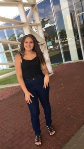 Imagem da aluna Larissa. Ela possui cabelos castanhos cacheados, calças jeans, uma blusa preta de alça e sandálias pretas.