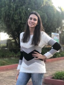 Foto de Kayza em uma rua arborizada. Ela tem cabelos castanhos compridos, usa calça jeans e uma blusa branca com listras cinzas e pretas de mangas compridas.