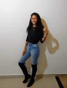Foto de Luana em frente a uma parede branca. Ela tem cabelos negros cacheados e usa calça jeans azul clara, uma blusa preta de manga na metade do braço e botas de cano alto pretas. 