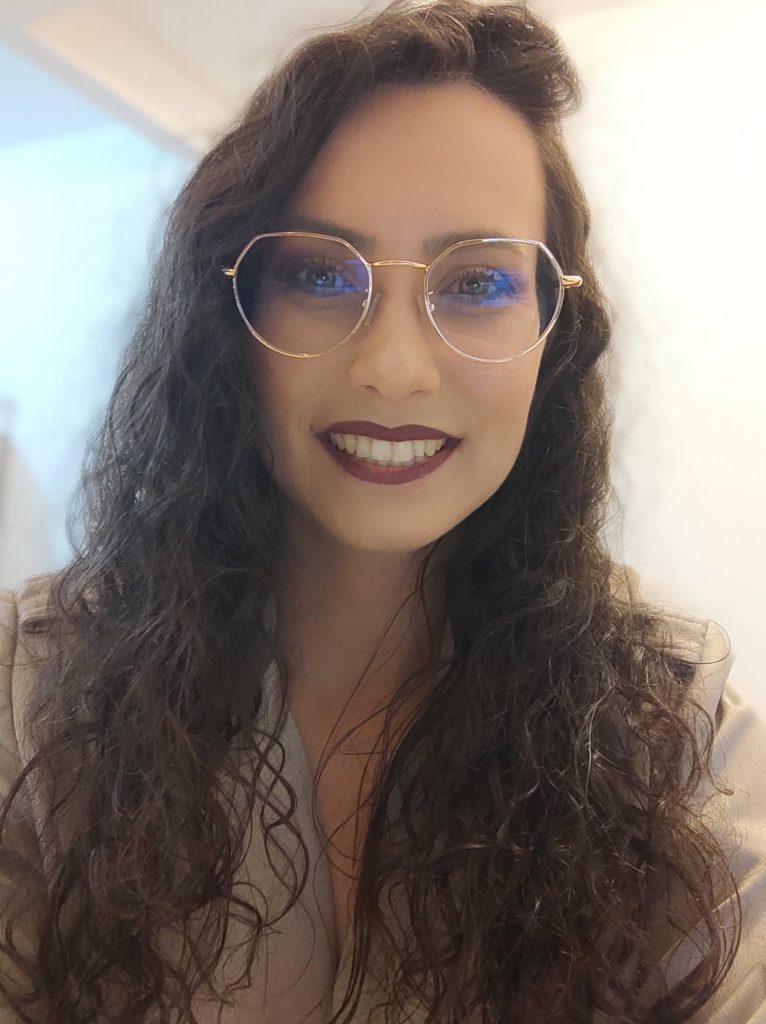 Foto de Kyene Becker. Ela é uma mulher branca de cabelos castanhos compridos e cacheados. Usa óculos de grau redondo e batom roxo na boca, que sorri.