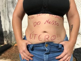 Foto de uma pessoa, aparencendo a barriga de fora, escrito "tire suas regras do nosso útero". A pessoa está com a mão no coz da calça e está de top. Não aparece seu rosto.