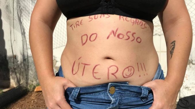 Foto de uma pessoa, aparencendo a barriga de fora, escrito "tire suas regras do nosso útero". A pessoa está com a mão no coz da calça e está de top. Não aparece seu rosto.