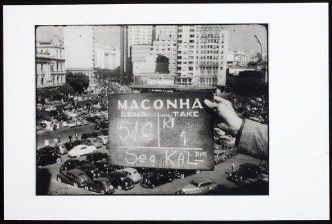 Reprodução de um frame cinematográfico em preto e branco, onde há uma pessoa segurando uma placa onde lê-se "Maconha Cena 51C Take 1 Seg Kal". Atrás há uma estacionamento de carros da década de 40 com prédios ao redor.