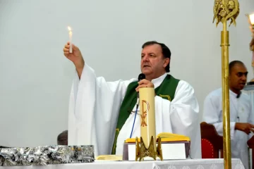 Retrato do Padre Ticão durante cerimônia religiosa, com vestes sacerdotais, segurando uma vela. Ele é um homem de meia idade, de pele bege claro, com cabelos pretos levemente calvos. Suas vestes são brancas e compridas, sobre as quais há um uma faixa verde escuro com bordados dourados. Ele fala ao microfone, em frente a uma mesa de sua igreja, onde há objetos religiosos como uma bíblia aberta, vela e decorações.