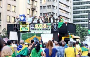 Foto com uma estrutura de metal, formando um palanque, com pessoas falando ao microfone e, abaixo, de costas, várias pessoas escutando. Todas elas vestidas com as cores da bandeira brasileira. Há placas escrito "s.o.s. forças armadas" e "intervenção militar já"